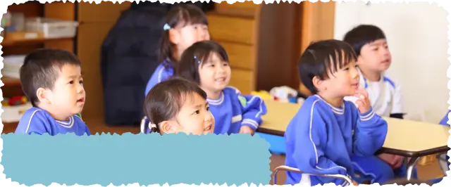 青い体操服を着た園児たちが保育室で椅子に座り、先生の話を笑顔で聞いている様子