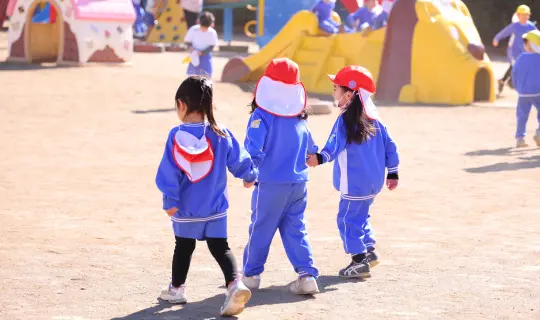 園庭で手をつなぎ歩く園児3人の後ろ姿